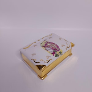 May include: A white porcelain box shaped like a book, with gold trim and accents. The top features a cross design with purple and green floral elements. The box has a rectangular shape.