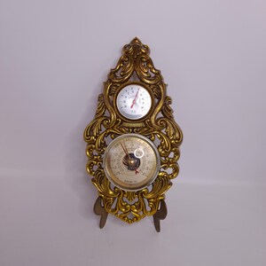 May include: A vintage gold-colored barometer and thermometer combination. The ornate frame features scrolling details. The thermometer is at the top, and the barometer is below. The barometer has a needle and a dial with text.