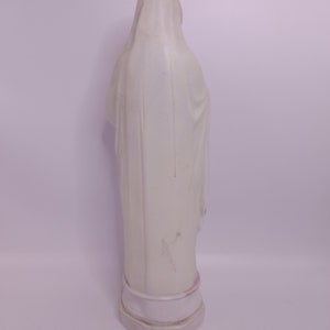 Puede incluir: Estatua de cer&aacute;mica blanca de una mujer con una t&uacute;nica larga. La estatua est&aacute; de pie sobre una base blanca.