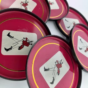 Op de afbeelding: Set van vintage rode metalen trays met zwarte randen en gouden accenten. Elke tray heeft een wit ruitvormig grafisch ontwerp van een persoon in een rode jas en zwarte laarzen. De trays zijn in een cirkelvorm gerangschikt.