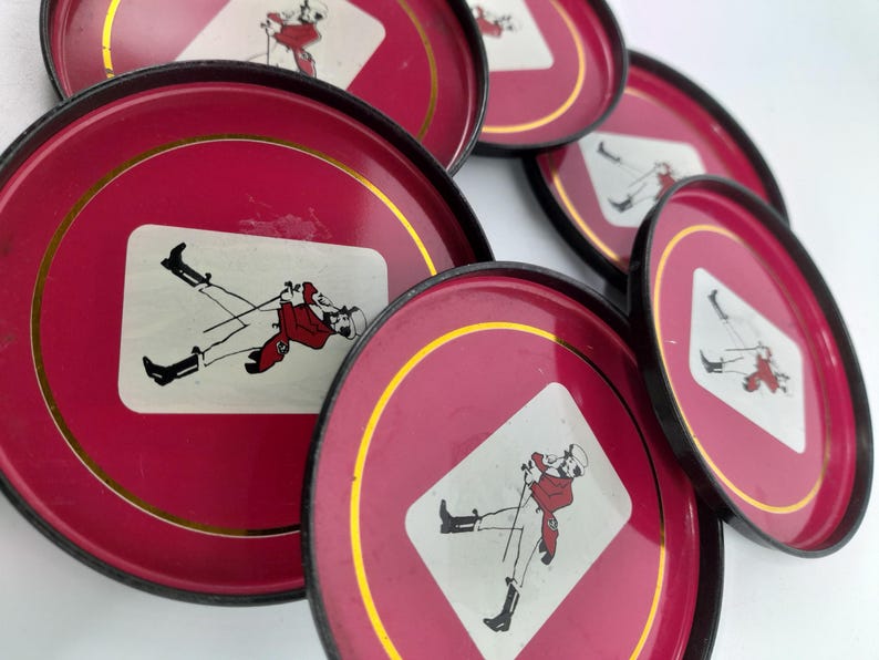 Op de afbeelding: Set van vintage rode metalen trays met zwarte randen en gouden accenten. Elke tray heeft een wit ruitvormig grafisch ontwerp van een persoon in een rode jas en zwarte laarzen. De trays zijn in een cirkelvorm gerangschikt.