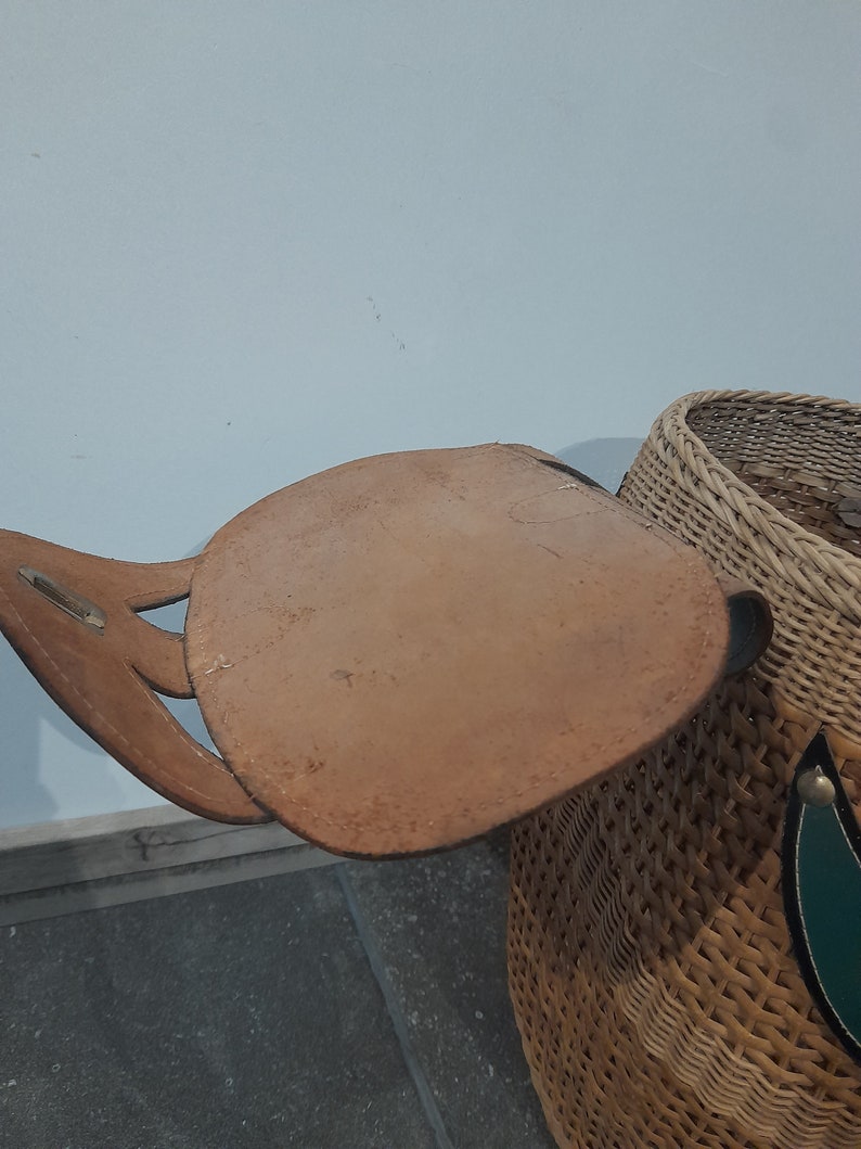 May include: A woven wicker basket with a brown leather strap and handle. The basket has a green and white design on the side.