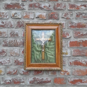 Könnte beinhalten: Ein gerahmtes religiöses Kruzifix mit einer silbernen Christusfigur auf einem Holzkreuz. Das Kreuz wird vor einem grünen Stoffhintergrund in einem dekorativen goldfarbenen Rahmen präsentiert. Der Rahmen ist an einer verwitterten Backsteinmauer befestigt.