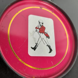 Op de afbeelding: Rode blikken doos met een gouden rand en een wit etiket met een figuur in een rode jas en zwarte laarzen. De doos is omgeven door andere zwarte blikken, wat op een set duidt.