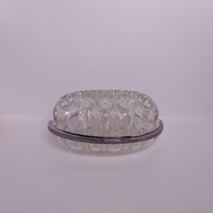 May include: A clear glass butter dish with a decorative, textured design. The dish has a round shape with a lid and a dark grey band around the base. The design features a series of rounded shapes.