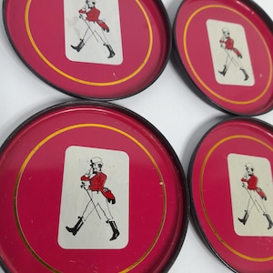 Op de afbeelding: Set van vier rode metalen onderzetters met een gouden rand en een zwarte rand. Elke onderzetter heeft een witte rechthoek met een afbeelding van een man in een rode jas en zwarte laarzen. De onderzetters zijn rond.