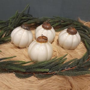 Op de afbeelding: Vier witte glazen kerstballen met een robuuste bruine metalen bovenkant. De ballen liggen op een bruin nep bont oppervlak met een groene krans eromheen.