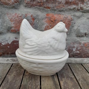 May include: A cream-colored ceramic egg keeper featuring a lid shaped like a sitting hen with detailed feathers and a base designed to resemble a woven basket. The two-piece container has a smooth, glossy finish and a rounded, oval shape.