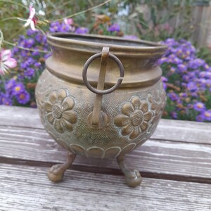Pu&ograve; includere: Un vaso vintage in ottone con manico e tre piedini a zampa. Il vaso presenta disegni floreali in rilievo attorno al corpo. Lo sfondo include una superficie in legno e fiori sfocati.