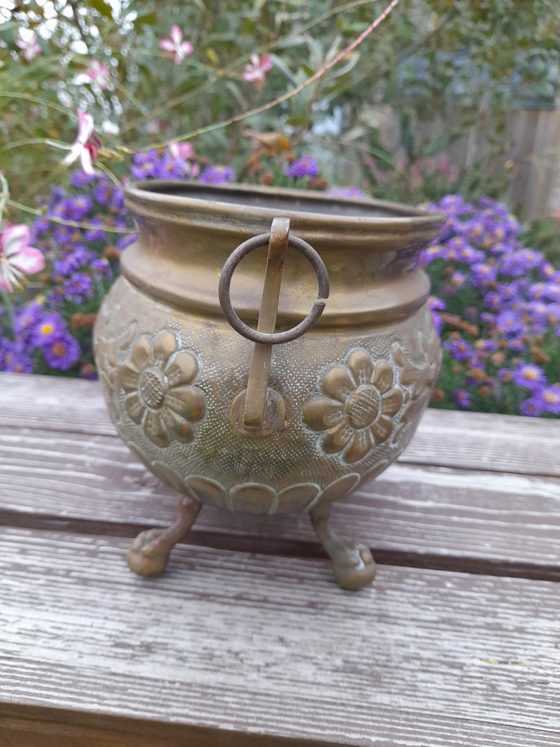 Pu&ograve; includere: Un vaso vintage in ottone con manico e tre piedini a zampa. Il vaso presenta disegni floreali in rilievo attorno al corpo. Lo sfondo include una superficie in legno e fiori sfocati.