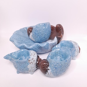 Op de afbeelding: Een set van vier keramische schelpvormige kommen en bakjes met een lichtblauwe glazuurlaag, donkerblauwe wervelpatronen en bruine accenten. De stukken hebben een textuurafwerking en zijn gegroepeerd tegen een witte achtergrond.