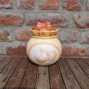May include: A ceramic jar with a lid, featuring a design of two garlic bulbs. The jar is cream-colored with a peach-colored border and a decorative bow on the lid. The jar is sitting on a wooden surface.