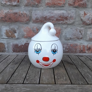 May include: A white ceramic jar with a clown face design. The jar has a round body, a curved lid, and is decorated with blue eyes, a red nose, and a red smiling mouth. The jar is sitting on a wooden surface.