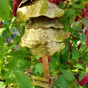 May include: A gold metal wind chime with multiple layers of hammered metal discs hanging from a copper pipe. The chime is attached to a green plant with red flowers.