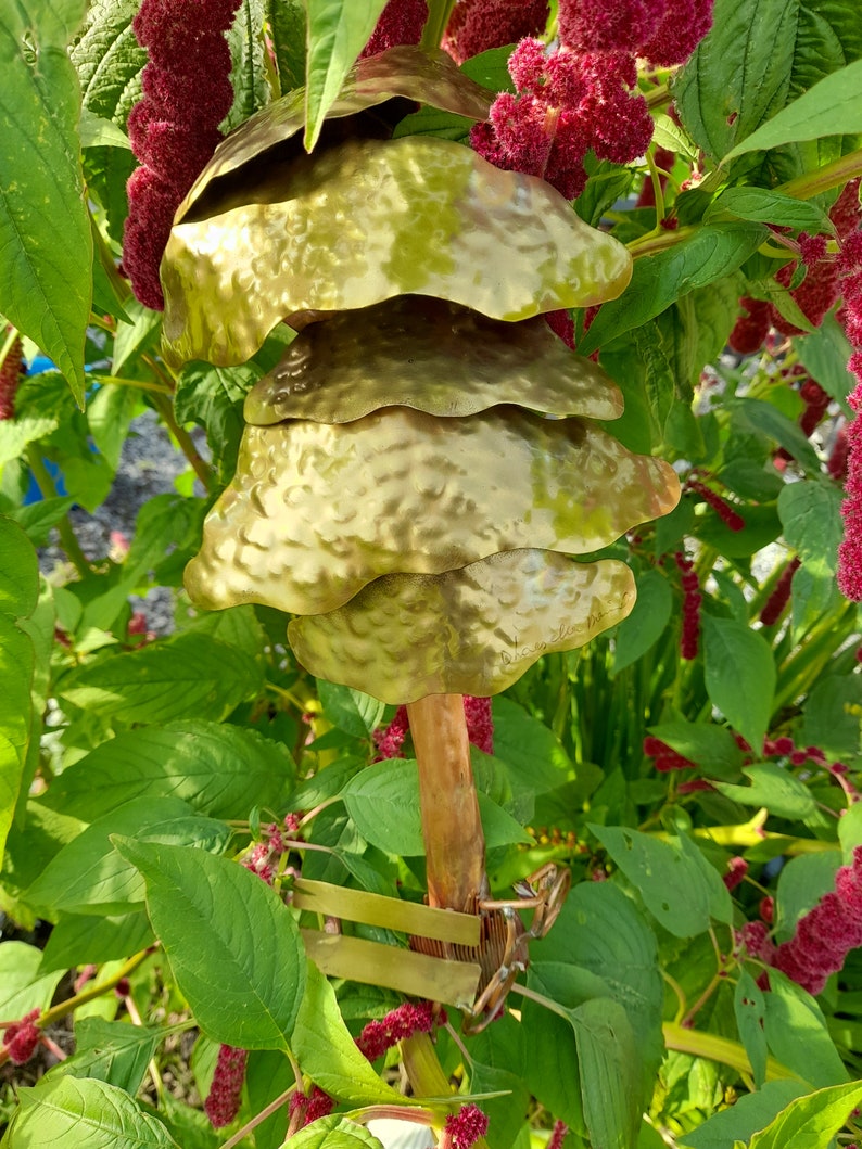 May include: A gold metal wind chime with multiple layers of hammered metal discs hanging from a copper pipe. The chime is attached to a green plant with red flowers.