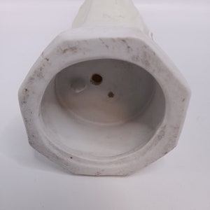 Puede incluir: Jarr&oacute;n de cer&aacute;mica blanco con base hexagonal y parte superior lisa y redondeada. El jarr&oacute;n tiene dos peque&ntilde;os agujeros en la parte inferior.