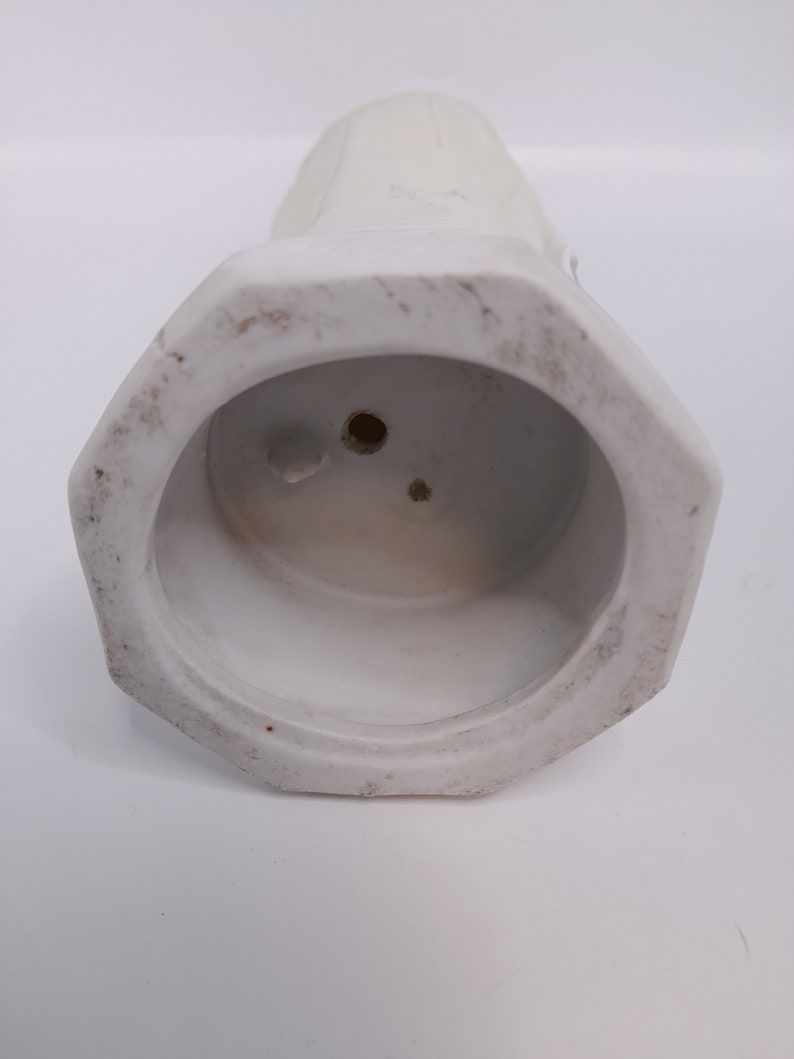 Puede incluir: Jarr&oacute;n de cer&aacute;mica blanco con base hexagonal y parte superior lisa y redondeada. El jarr&oacute;n tiene dos peque&ntilde;os agujeros en la parte inferior.