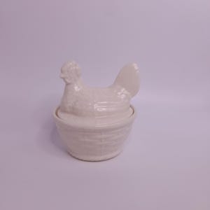 Op de afbeelding: Een witte keramische container in de vorm van een kip met een deksel. De kip zit op een mandachtige basis. De container wordt waarschijnlijk gebruikt voor het bewaren van eieren of andere kleine items. Het ontwerp is eenvoudig en elegant.