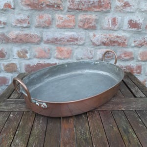 May include: An oval-shaped copper roasting pan with a metallic interior lining. The pan features two curved copper handles attached to the sides with rivets, providing a sturdy grip for lifting and moving the cookware during use.
