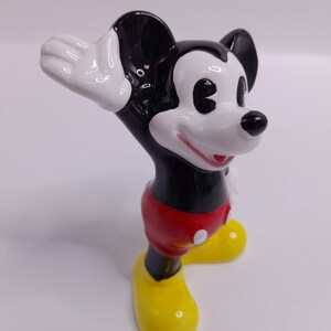 May include: A ceramic figurine of Mickey Mouse with black and white fur, red shorts, and yellow shoes. He is waving with his right hand.
