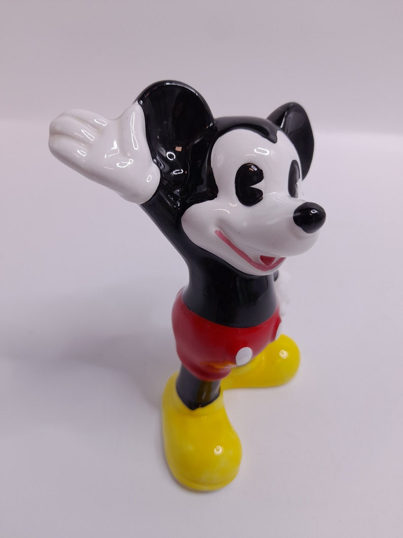 May include: A ceramic figurine of Mickey Mouse with black and white fur, red shorts, and yellow shoes. He is waving with his right hand.