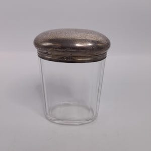 May include: A clear glass jar with a silver-colored lid. The lid has a textured surface and fits snugly on the jar. The jar is cylindrical with vertical ridges and is likely used for storing small items.