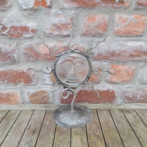 Puede incluir: Escultura decorativa de metal con forma de sol. El sol tiene una cara de cristal transparente con rasgos grabados y rayos metálicos. Se apoya sobre una base circular con un tallo curvo. Fondo: pared de ladrillo envejecida y superficie de madera.