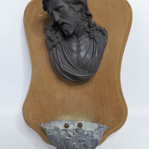K&ouml;nnte beinhalten: Eine Holztafel mit einer schwarzen Gipsb&uuml;ste von Jesus, der eine Dornenkrone tr&auml;gt. Ein kleines Metallbecken ist an der Unterseite der Tafel befestigt.