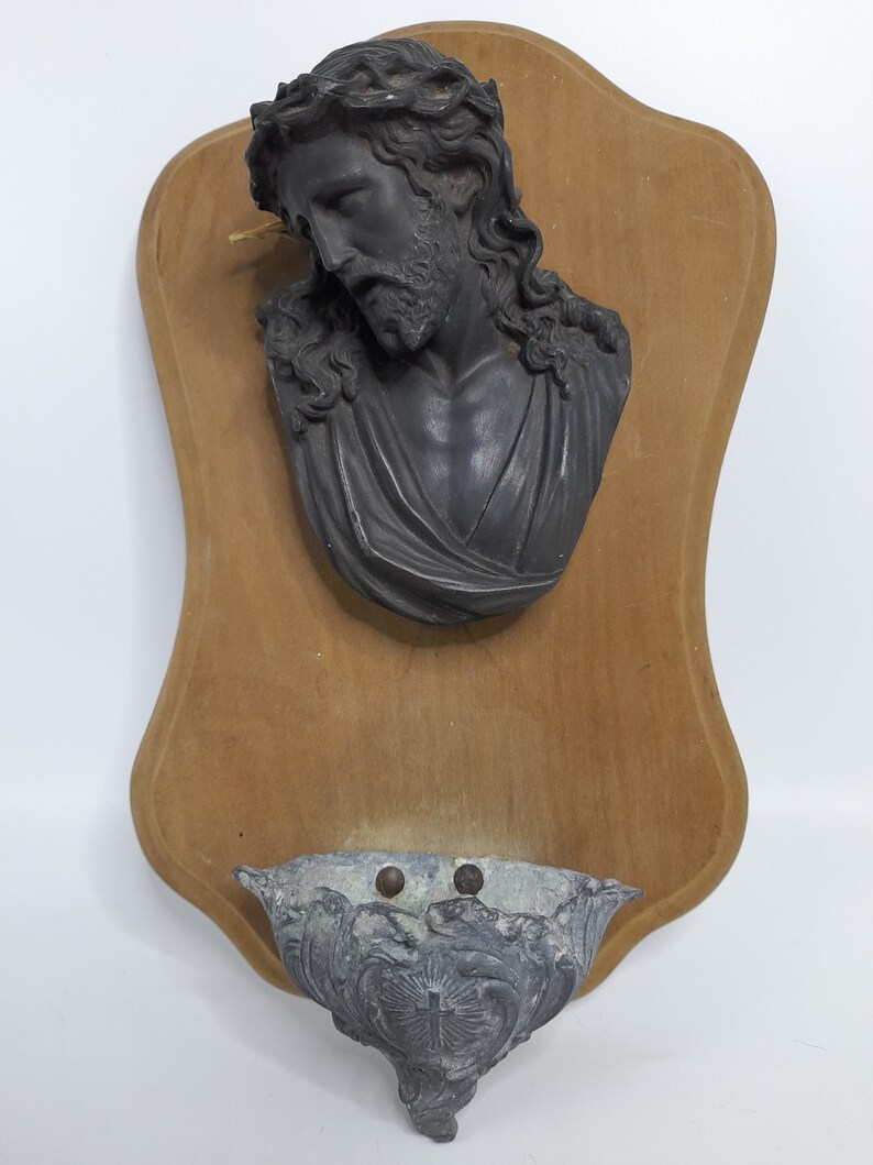 K&ouml;nnte beinhalten: Eine Holztafel mit einer schwarzen Gipsb&uuml;ste von Jesus, der eine Dornenkrone tr&auml;gt. Ein kleines Metallbecken ist an der Unterseite der Tafel befestigt.