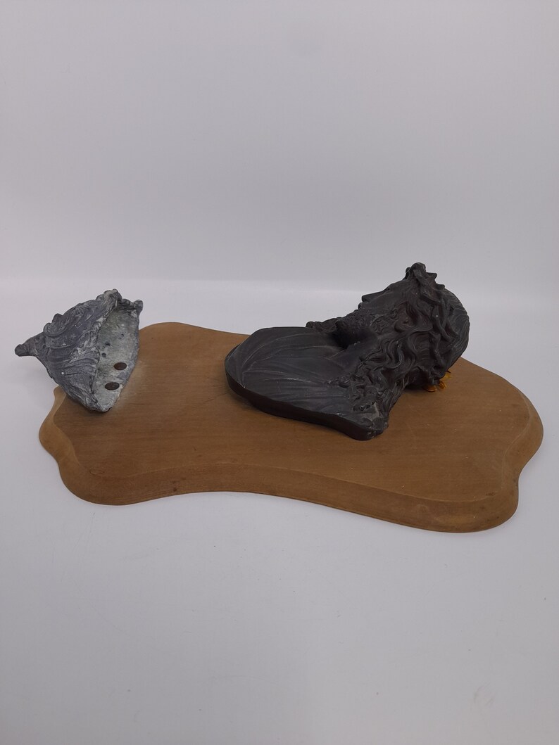 K&ouml;nnte beinhalten: Eine Holzplakette mit einer schwarzen und grauen Skulptur eines M&auml;nnerkopfes mit Dornenkrone. Eine graue und silberne Skulptur einer Muschel befindet sich ebenfalls auf der Plakette.