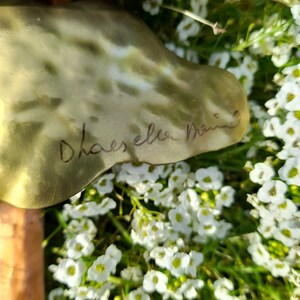 May include: A close-up of a gold metal sculpture with a signature that reads "Dhaeselen Design". The sculpture is surrounded by white flowers.
