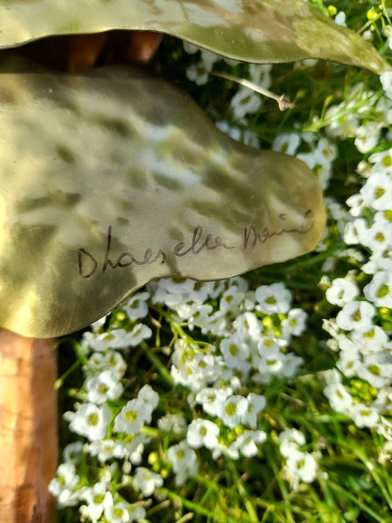 May include: A close-up of a gold metal sculpture with a signature that reads "Dhaeselen Design". The sculpture is surrounded by white flowers.