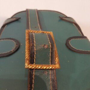 May include: Close-up of a green leather bag with a gold buckle. The buckle is rectangular with a rope-like design around the edge.