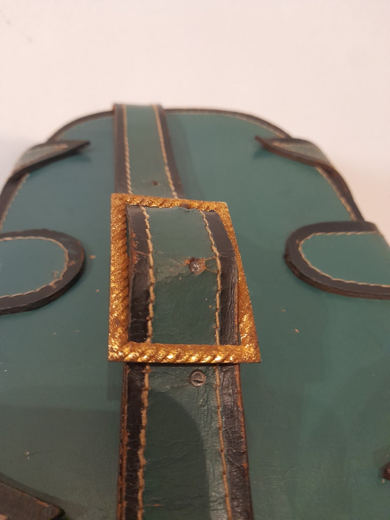 May include: Close-up of a green leather bag with a gold buckle. The buckle is rectangular with a rope-like design around the edge.