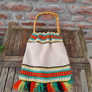 Pode incluir: Uma bolsa tote bege com alça de bambu. A bolsa apresenta padrões tecidos coloridos em vermelho, verde, amarelo e preto. A borda inferior tem uma franja de fios vermelhos, amarelos, verdes e pretos.