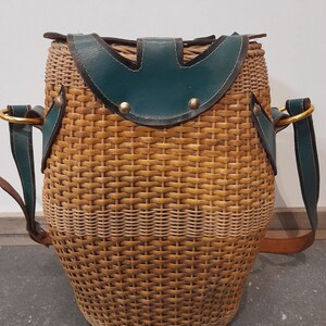 May include: A woven wicker bag with a green leather flap and brown leather straps. The bag has a round shape and a woven pattern.