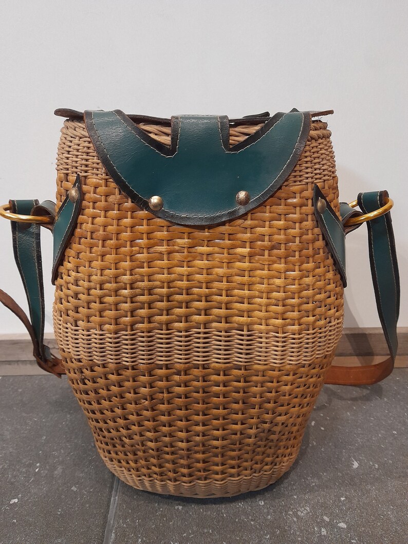 May include: A woven wicker bag with a green leather flap and brown leather straps. The bag has a round shape and a woven pattern.