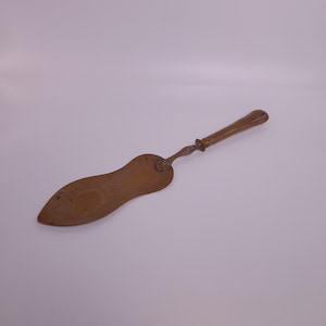 Könnte beinhalten: Ein Kuchenservierer im Vintage-Stil mit einem langen, verzierten Griff und einer breiten, gebogenen Klinge. Der Servierer ist aus warmem, goldbraunem Metall gefertigt und weist dekorative Details am Griff und an der Klinge auf. Das Design deutet auf eine klassische, antike Ästhetik hin.