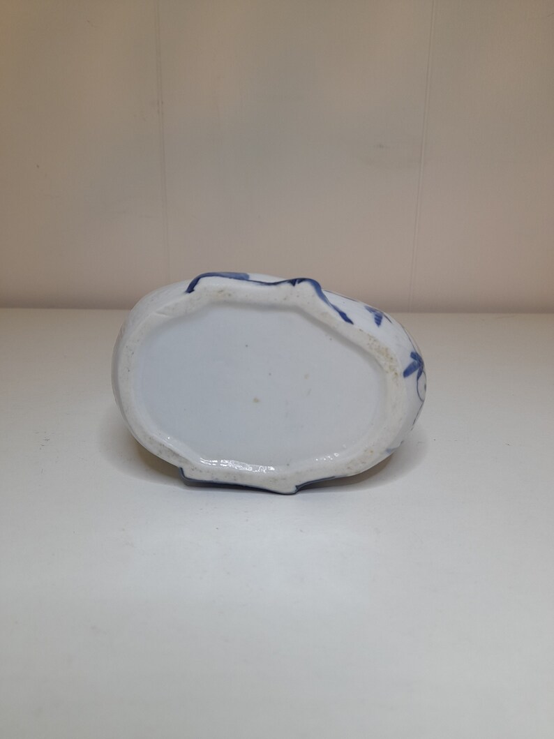 May include: The bottom of a white ceramic vase with blue floral designs. The vase has a rounded shape and a slightly raised rim.