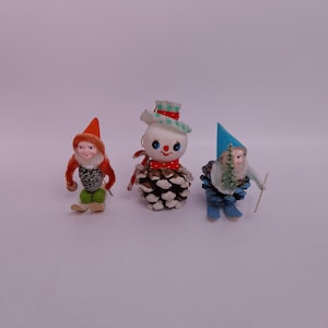Peut inclure: Trois ornements de Noël vintage. Un petit gnome avec un chapeau orange et des skis verts, un bonhomme de neige avec un corps en pomme de pin et un gnome avec un chapeau bleu et des skis. Les ornements sont sur fond blanc.