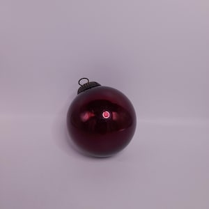 May include: A single, round, burgundy Christmas bauble with a shiny, reflective surface. The bauble has a small, dark metal cap with a loop for hanging. The background is a plain, light purple.