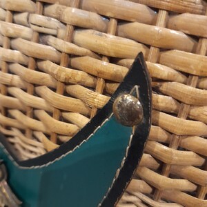 May include: Close-up of a woven wicker basket with a teal leather strap and a silver metal buckle. The strap has black stitching around the edges.