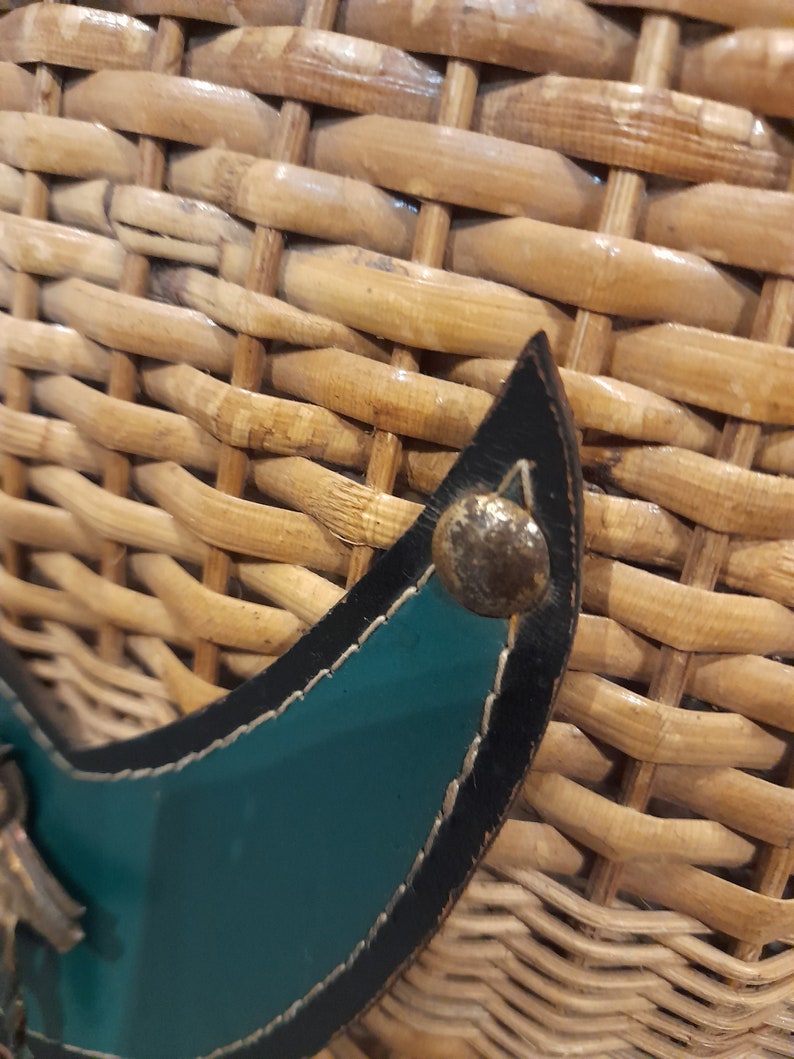 May include: Close-up of a woven wicker basket with a teal leather strap and a silver metal buckle. The strap has black stitching around the edges.