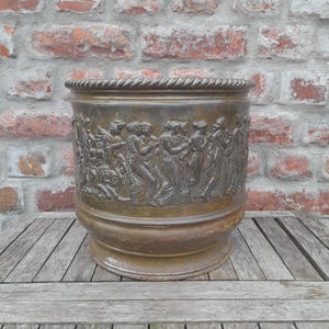 Puede incluir: Una maceta cilíndrica de bronce con un friso en relieve detallado de figuras clásicas con túnicas fluidas que rodea el exterior. El borde tiene un patrón de cuerda trenzada y la base es acampanada. El metal tiene un acabado de pátina oscura.
