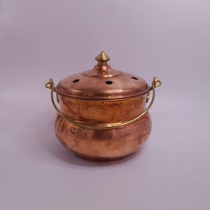 May include: A round copper pot with a brass swing handle and a matching copper lid. The lid features a central brass finial and several small circular ventilation holes arranged in a ring. The pot has a smooth, polished finish with visible metallic sheen.