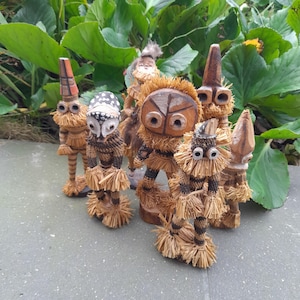 Op de afbeelding: Een groep van zeven houten beeldjes met strooiachtige haren en kleding. De beeldjes hebben geschilderde gezichten en staan op een grijs oppervlak.