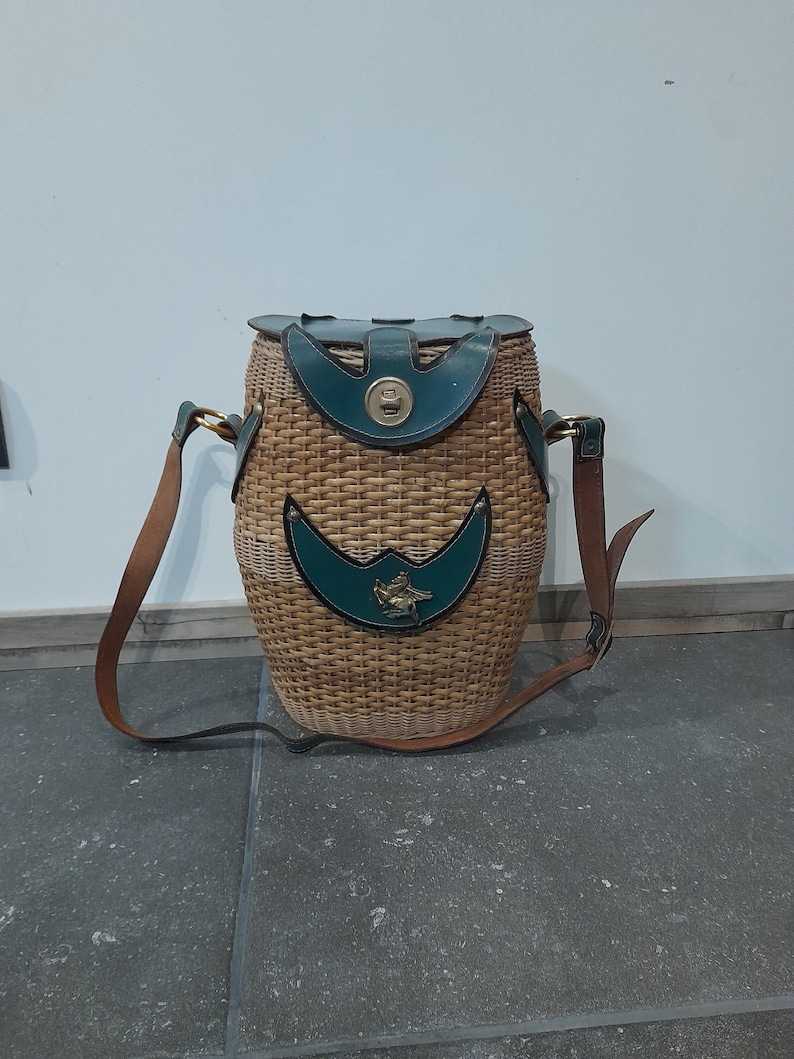 May include: A brown wicker basket with a green leather top and a brown leather strap. The basket has a brass buckle and a green leather patch with a gold design.