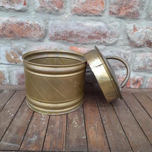 Pode incluir: Um recipiente redondo de latão com uma tampa. O recipiente tem um padrão decorativo canelado. A tampa tem uma alça.