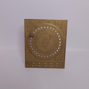 May include: A gold-colored, rectangular calendar with a circular dial featuring numbers 1-31. The calendar has a sun design in the center and Roman numerals along the bottom. Ornate scrollwork decorates the corners.