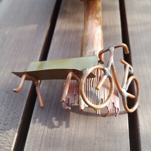 May include: A copper sculpture of a tree with a bench and a bicycle underneath. The sculpture is made of wire and has a rustic look.